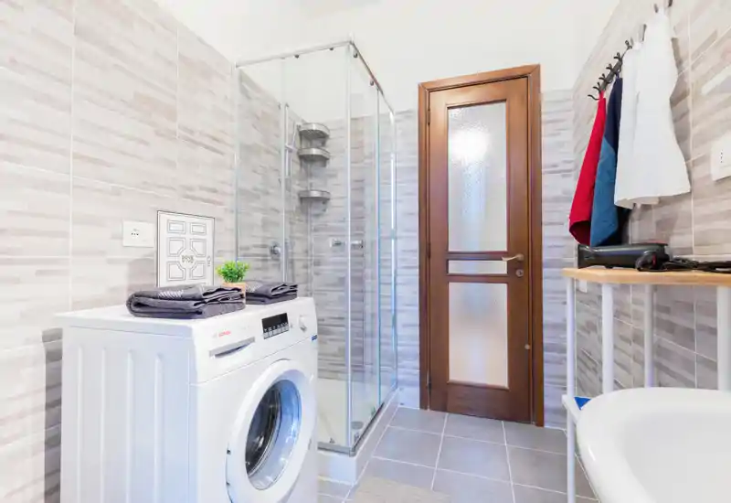 Bright, modern bathroom with a corner glass shower, washing machine and wooden door — neat tiles and practical storage create a clean, functional space.