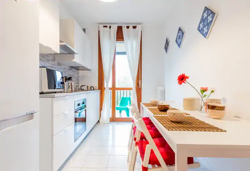 Bright, modern galley kitchen with white cabinetry, integrated appliances and a small dining counter with red-cushioned chairs; natural light enters through a glass door to a small balcony.