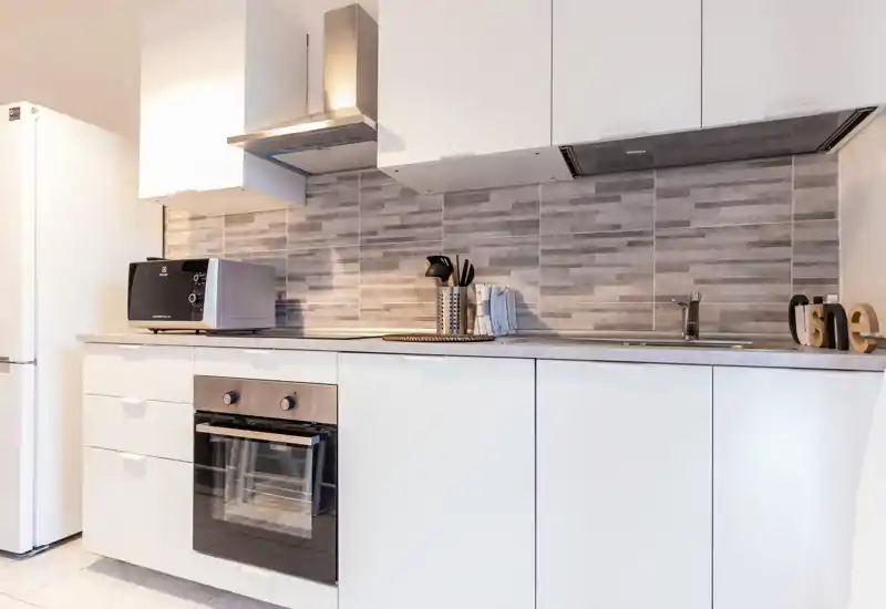 Modern compact kitchen with white cabinetry, built-in oven, stovetop hood and a neat tiled backsplash. Bright, minimal and well-organized space ideal for rental listings.