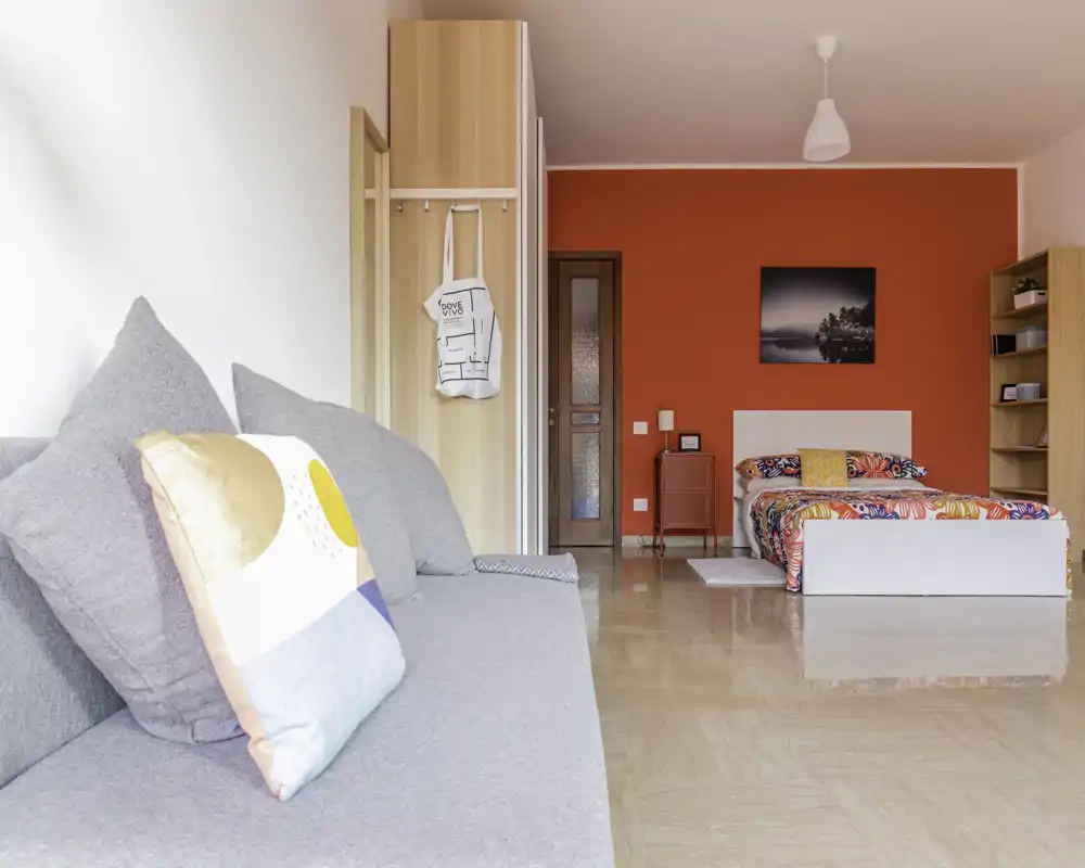 Bright, modern bedroom with a colorful bed against a bold red accent wall and a comfy grey sofa in the foreground. Large tiles and clean lines give a polished contemporary feel, ideal for rental listings highlighting space and style.
