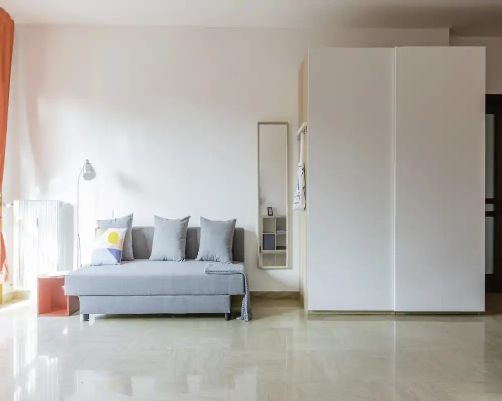 Bright, minimalist living area with a light grey sofa, decorative cushions and a large white wardrobe — modern, airy space with natural light.