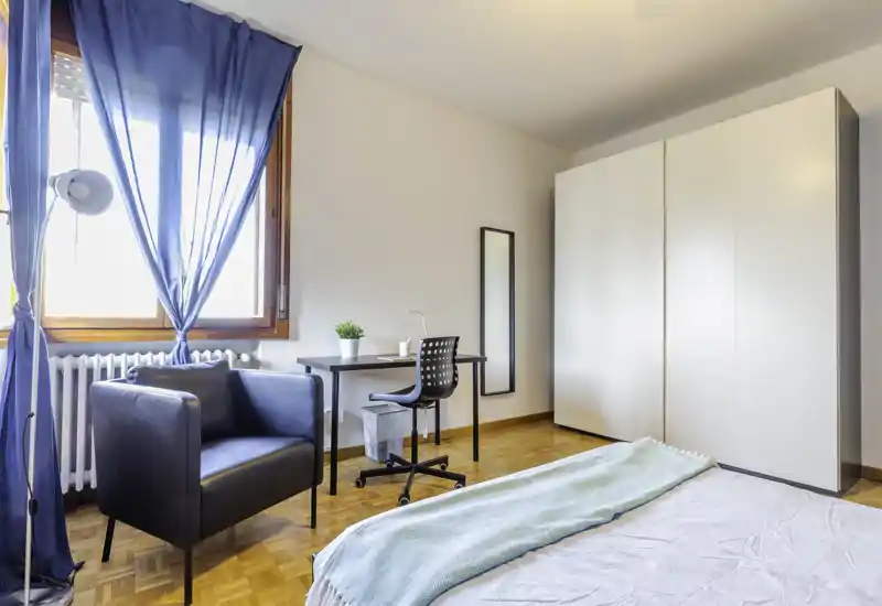 Camera da letto luminosa e moderna con letto parzialmente visibile, postazione di lavoro con scrivania e sedia, poltrona vicino alla finestra e grande armadio — ideale per chi cerca un alloggio funzionale e di stile.