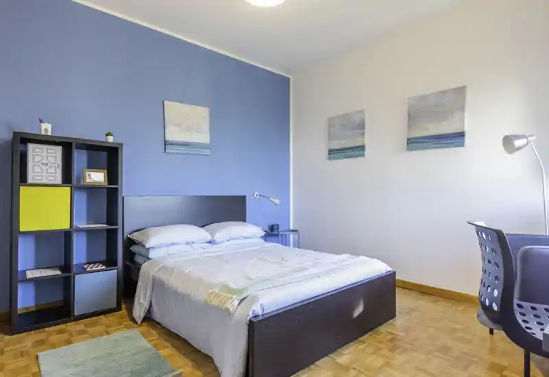Camera da letto luminosa e ben arredata con letto matrimoniale davanti a una parete blu, biancheria coordinata, comodino e libreria; pavimento in parquet e quadri dal tema marino creano un’atmosfera fresca e accogliente.