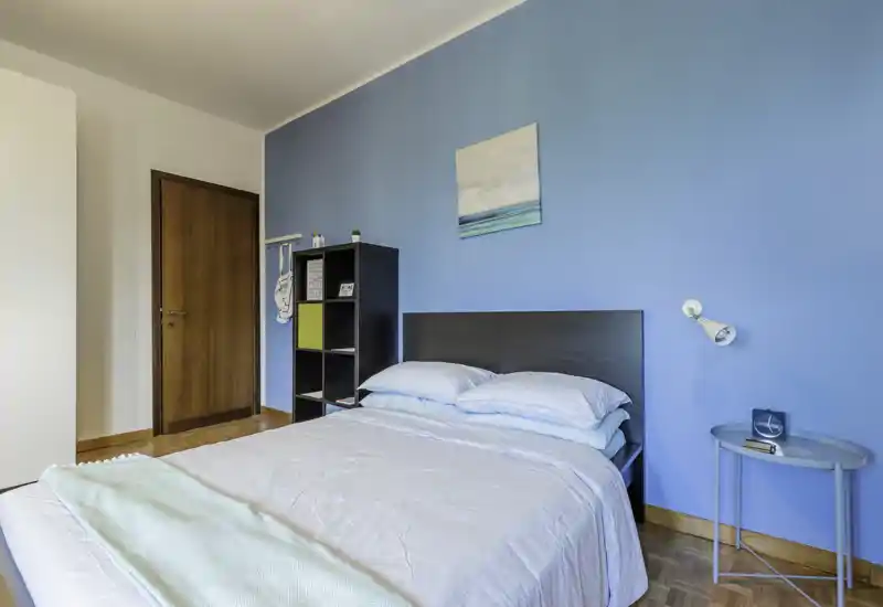 Camera da letto luminosa e ordinata con un letto matrimoniale, lenzuola morbide e una parete d'accento blu; scaffale semplice e piccolo comodino completano l'ambiente moderno e accogliente.