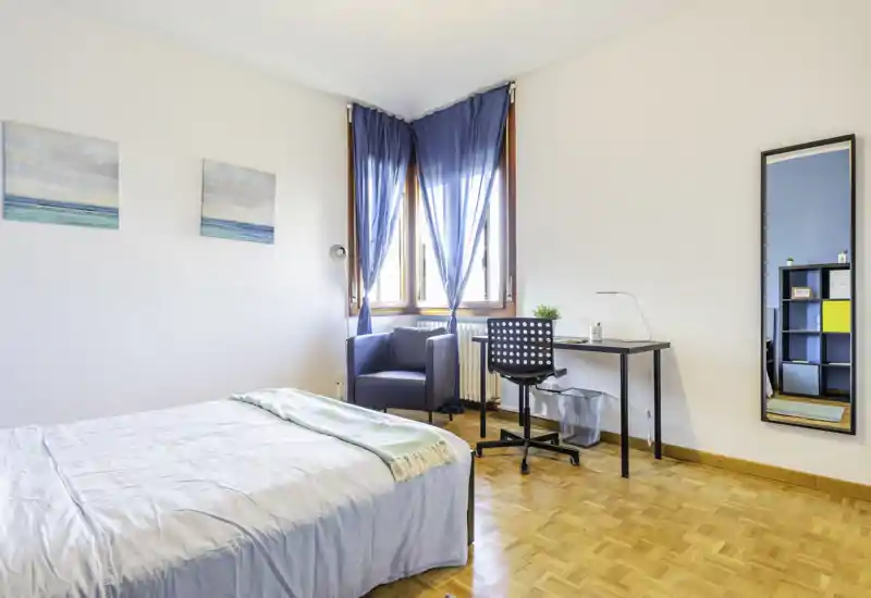 Camera da letto luminosa e ordinata con un comodo letto matrimoniale, scrivania vicino alla finestra e una poltrona accogliente — perfetta per riposare o lavorare.