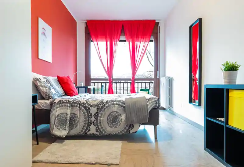 Bright, modern bedroom with patterned bedding, red accent wall and sheer red curtains framing a large window — cozy and well-styled for rental listings.