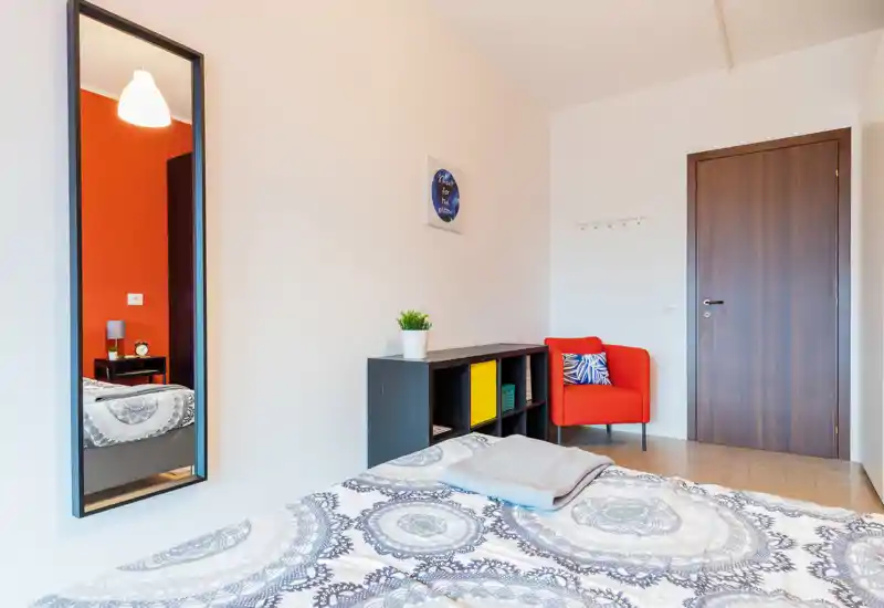 Bright, modern bedroom with patterned bedding, a full bed visible in the foreground, a small shelving unit and a red accent armchair near the door.