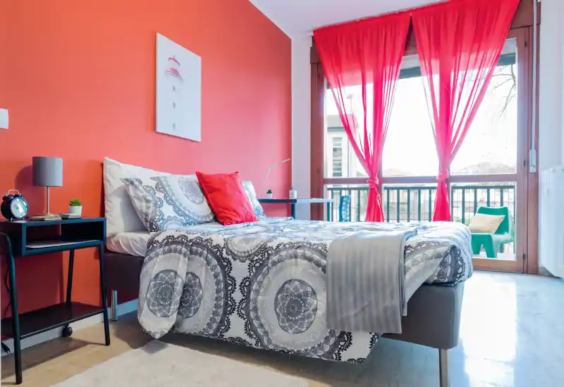 Bright, stylish bedroom with a large bed facing a balcony; warm red accent wall and coordinated textiles create a cozy feel, with abundant natural light.