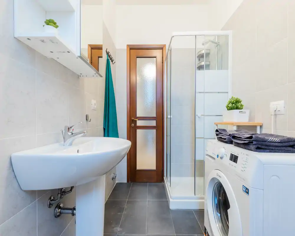 Bagno moderno e luminoso con lavabo a colonna, box doccia in vetro e una lavatrice a carica frontale posizionata sulla destra.