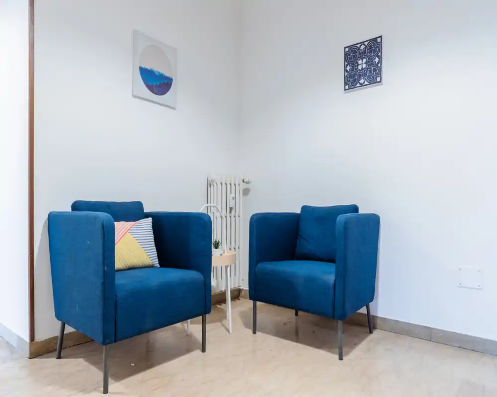 Angolo soggiorno con due poltrone blu, un cuscino decorativo e un tavolino laterale — luminoso, moderno e accogliente per rilassarsi o conversare.
