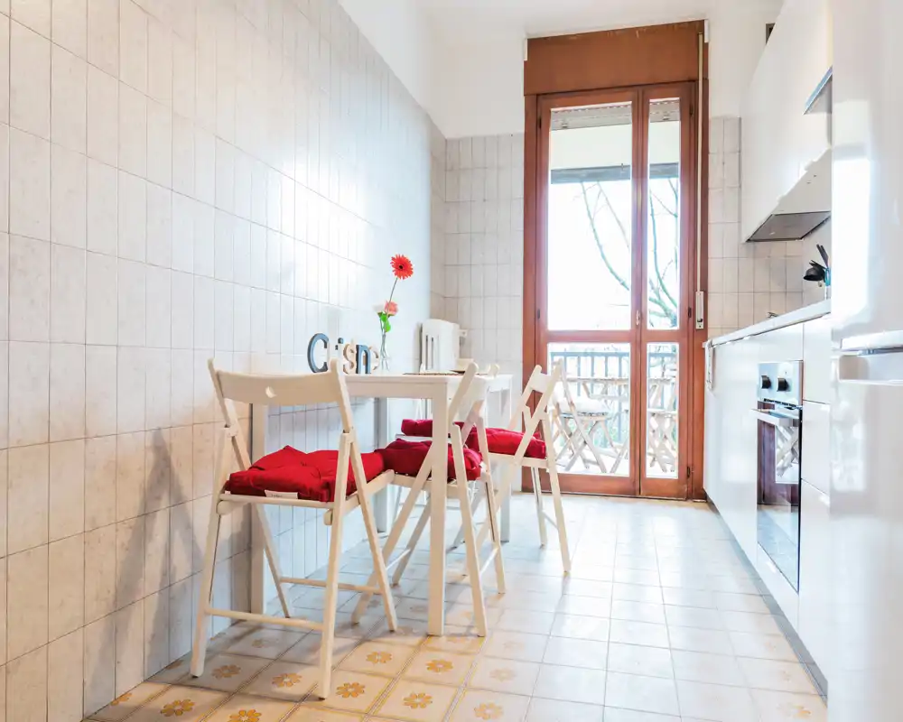 Cucina luminosa e compatta con mobili bianchi, forno integrato e un piccolo angolo pranzo con sedie pieghevoli e cuscini rossi. Porta finestra ampia verso il balcone che garantisce luce naturale e un angolo colazione accogliente.