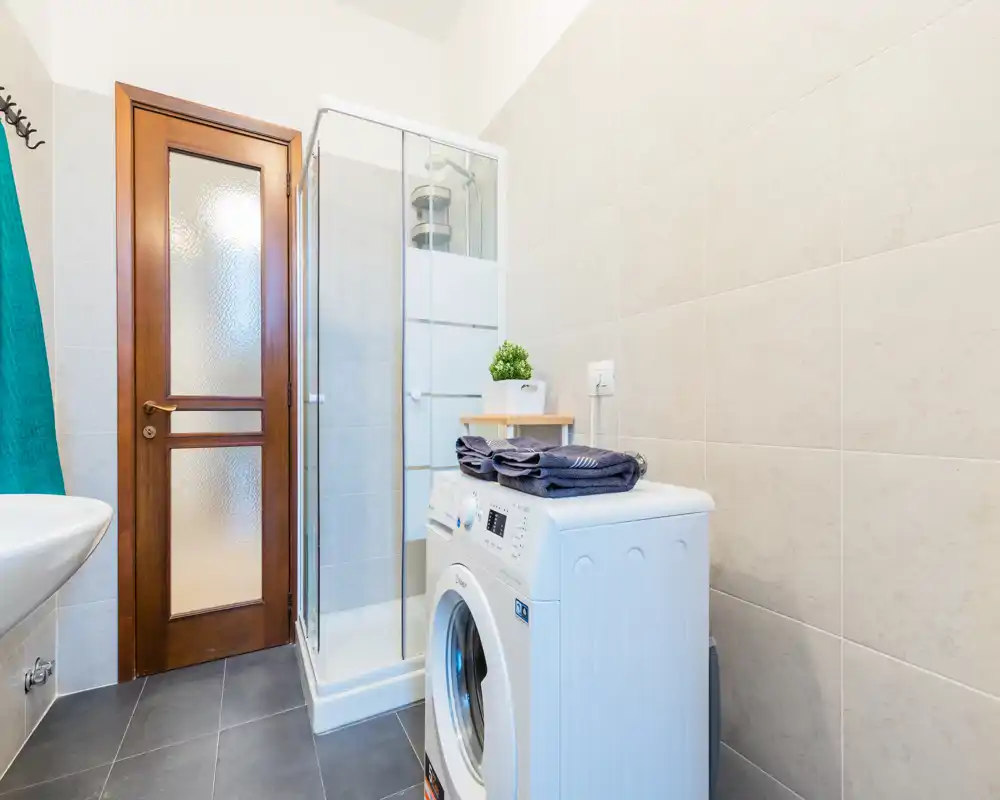 Bagno compatto e moderno con box doccia angolare, lavatrice e lavabo; piastrelle chiare e pavimento scuro offrono uno spazio pulito e funzionale.