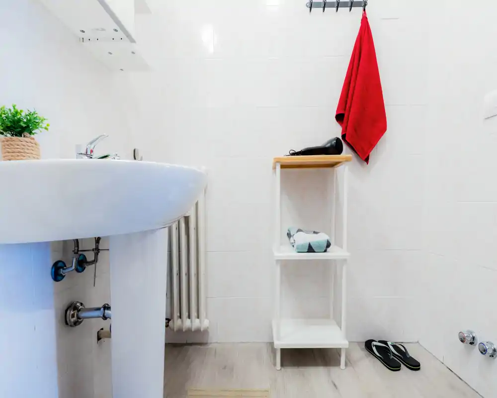 Bagno piccolo e luminoso con lavabo a colonna, piastrelle bianche e mensoline essenziali. Un asciugamano rosso e alcuni accessori danno un tocco di colore; il pavimento effetto legno completa l'aspetto pulito e minimal.