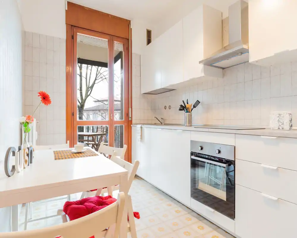 Cucina luminosa e moderna con mobili bianchi, forno incassato e piccolo tavolo da pranzo vicino alla porta del balcone che lascia entrare luce naturale — perfetta per colazioni accoglienti.