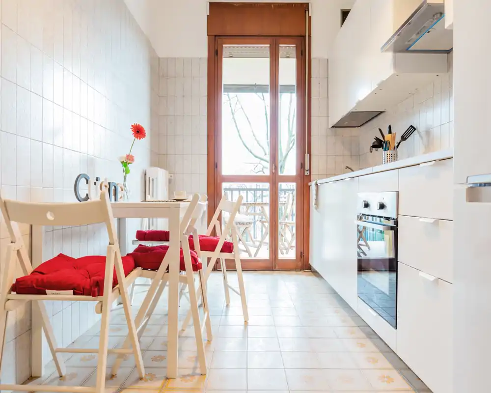 Cucina compatta e luminosa con mobili bianchi, piccolo angolo pranzo con sedie pieghevoli e cuscini rossi, porta finestra che illumina l'ambiente.