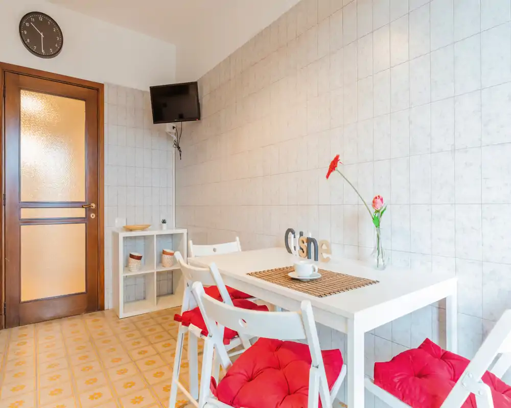 Zona pranzo luminosa e ordinata con tavolo bianco, sedie pieghevoli con cuscini rossi e un fiore decorativo — perfetta per pasti informali in un angolo accogliente e ben illuminato.