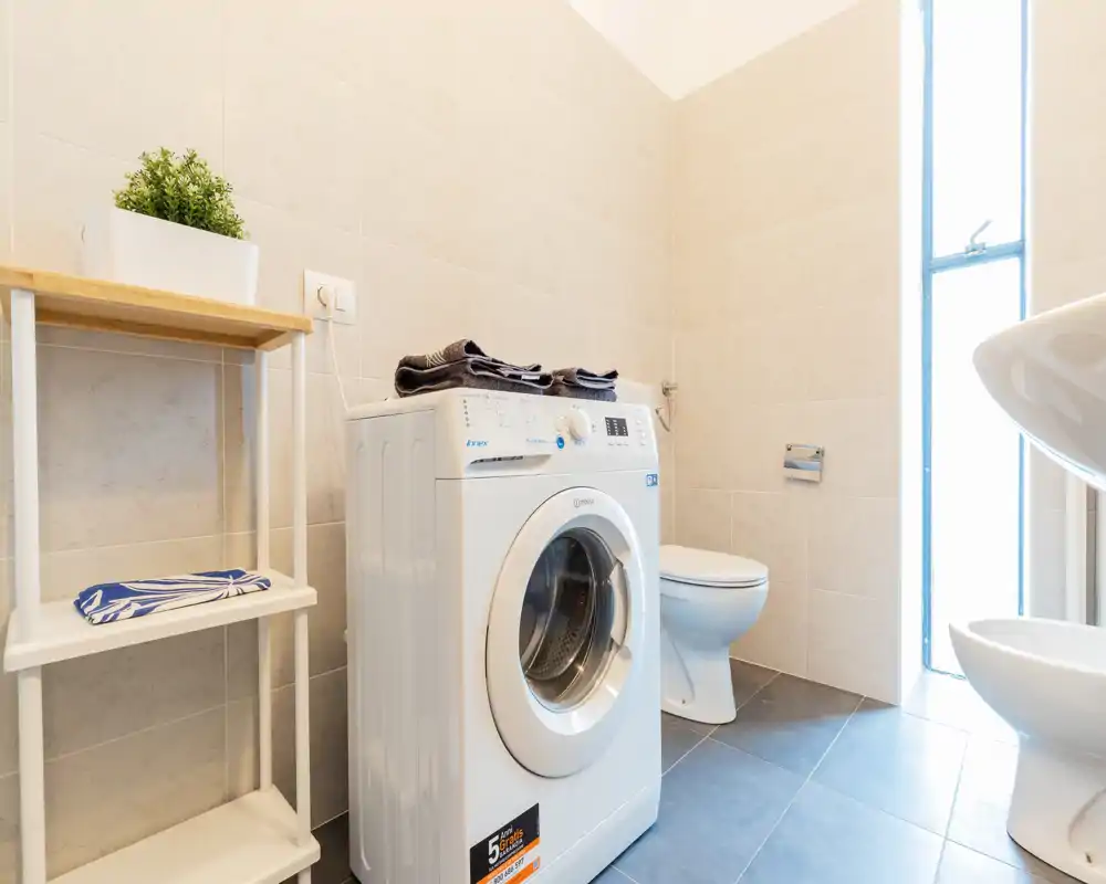 Bagno luminoso e moderno con lavatrice a carica frontale, WC e lavabo bianco. Piastrelle neutre e luce naturale creano un ambiente pulito e funzionale per bagno e lavanderia.