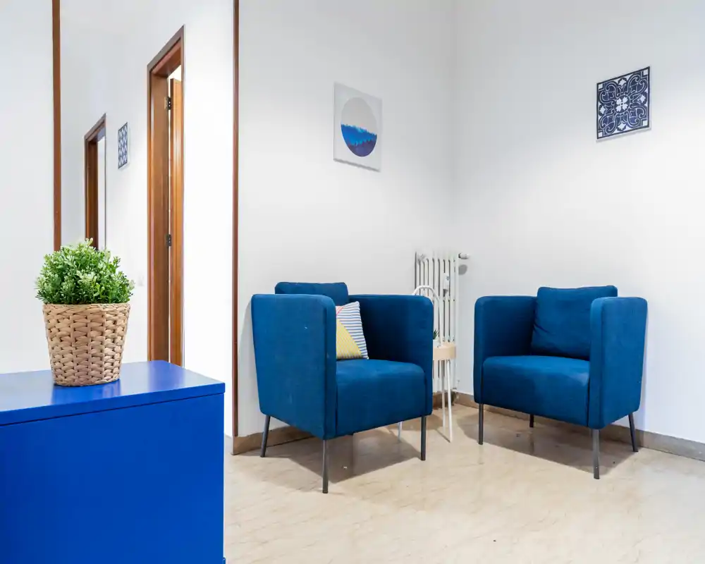 Angolo soggiorno luminoso e moderno con due poltrone blu, quadri semplici alle pareti e arredo minimal—ideale come zona relax o attesa.