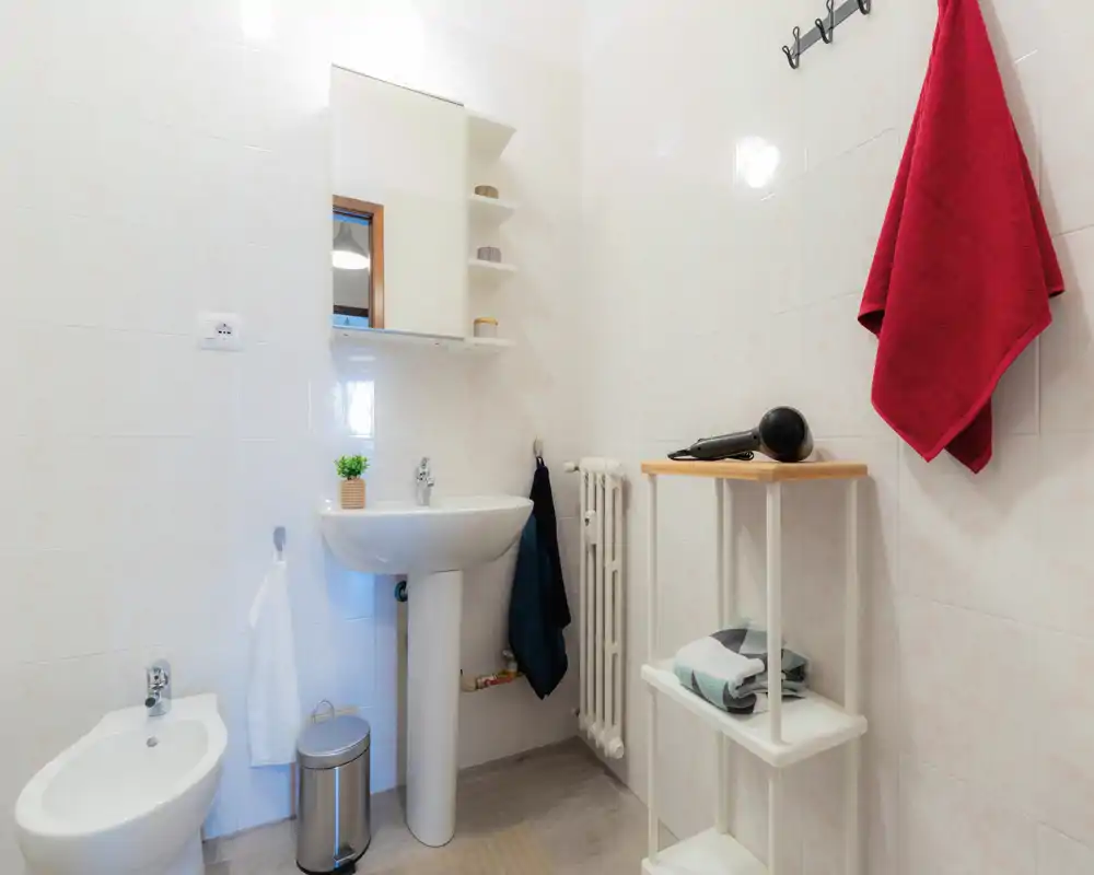 Bagno compatto e ordinato con lavabo a colonna, bidet, specchio con mobiletto e scaffalatura, ganci per asciugamani e piccolo mobiletto. Piastrelle chiare e accessori pratici adatti per affitti brevi.
