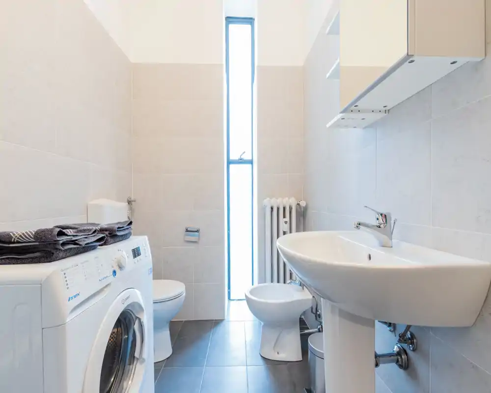 Bagno moderno e luminoso con lavabo a colonna, WC, bidet, termosifone e lavatrice, rivestimenti chiari e luce naturale proveniente da una finestra verticale.