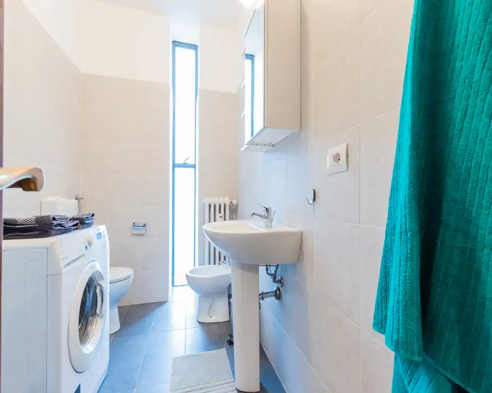 Bagno moderno e luminoso con lavabo a colonna, bidet, WC e lavatrice; pareti piastrellate e una finestra alta offrono luce naturale.