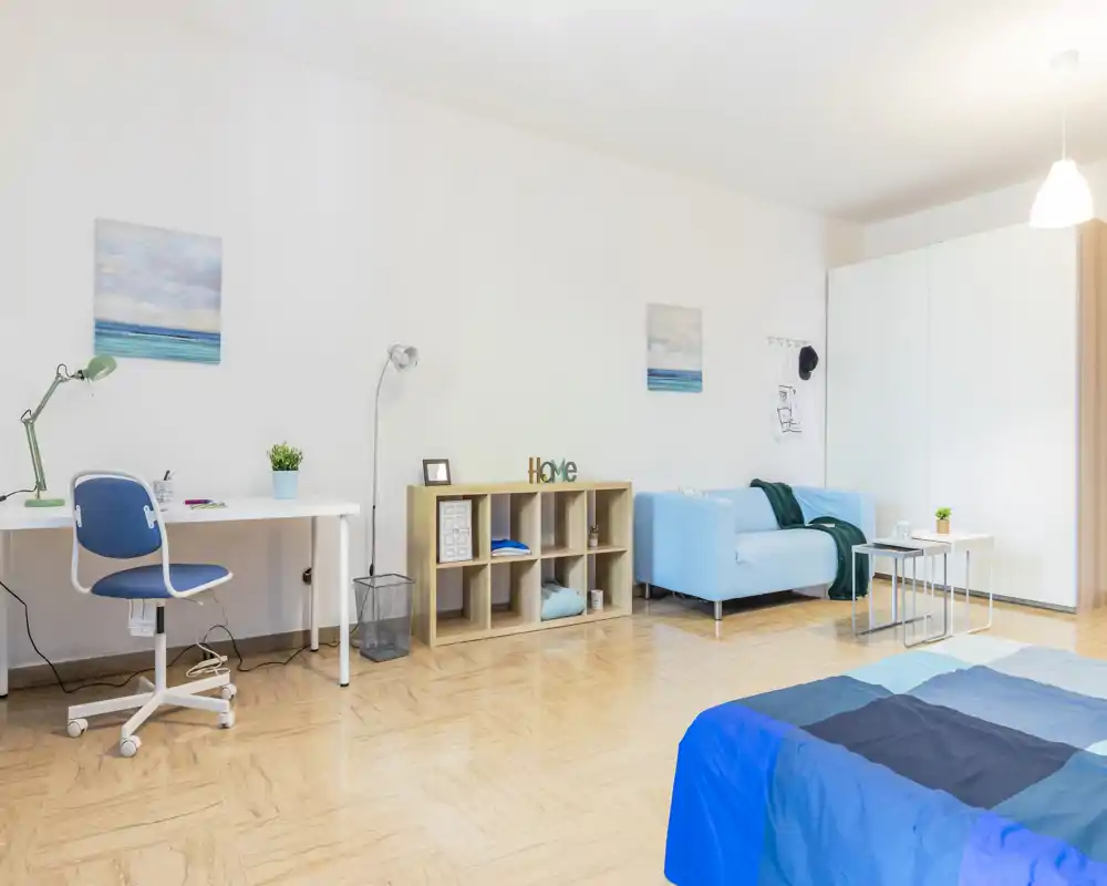Bright, tidy bedroom with a blue bedspread in the foreground, a small sofa and workspace along the wall, and clean minimalist décor—ideal for a single or student flat.