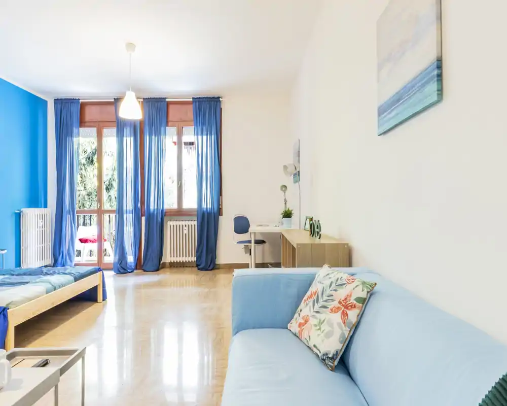 Spacious, bright bedroom with a light-blue sofa and wooden bed frame, large windows with blue curtains letting in abundant natural light and a tidy desk area — ideal for comfortable stays and work.