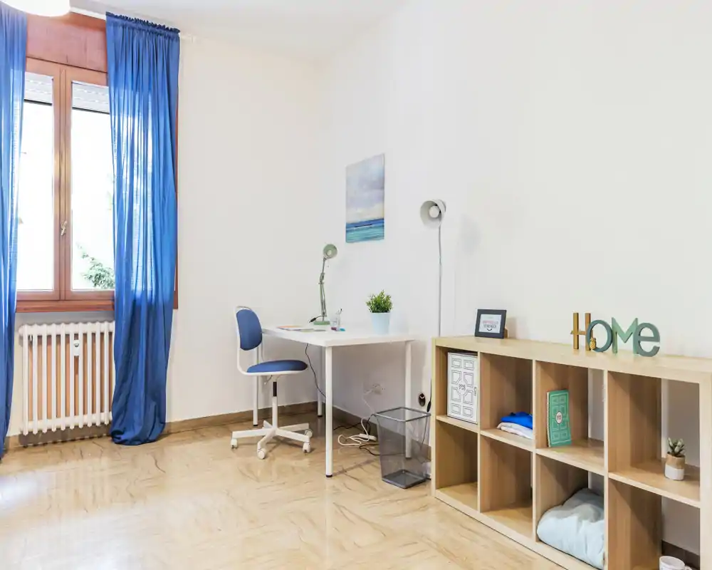 Bright, tidy living/study corner with a white desk, blue swivel chair and natural-wood shelving. The large window with blue curtains brings abundant daylight, and small decorative touches (plant, framed print) make the space welcoming.