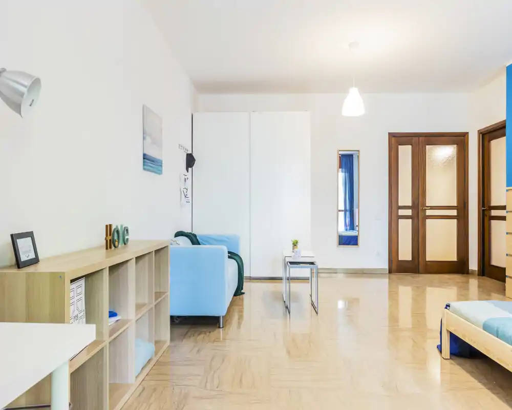 Bright, modern bedroom corner with a visible single bed frame at the right, a light-blue sofa, minimalist shelving and wooden doors. The space feels clean and airy with simple coastal accents.