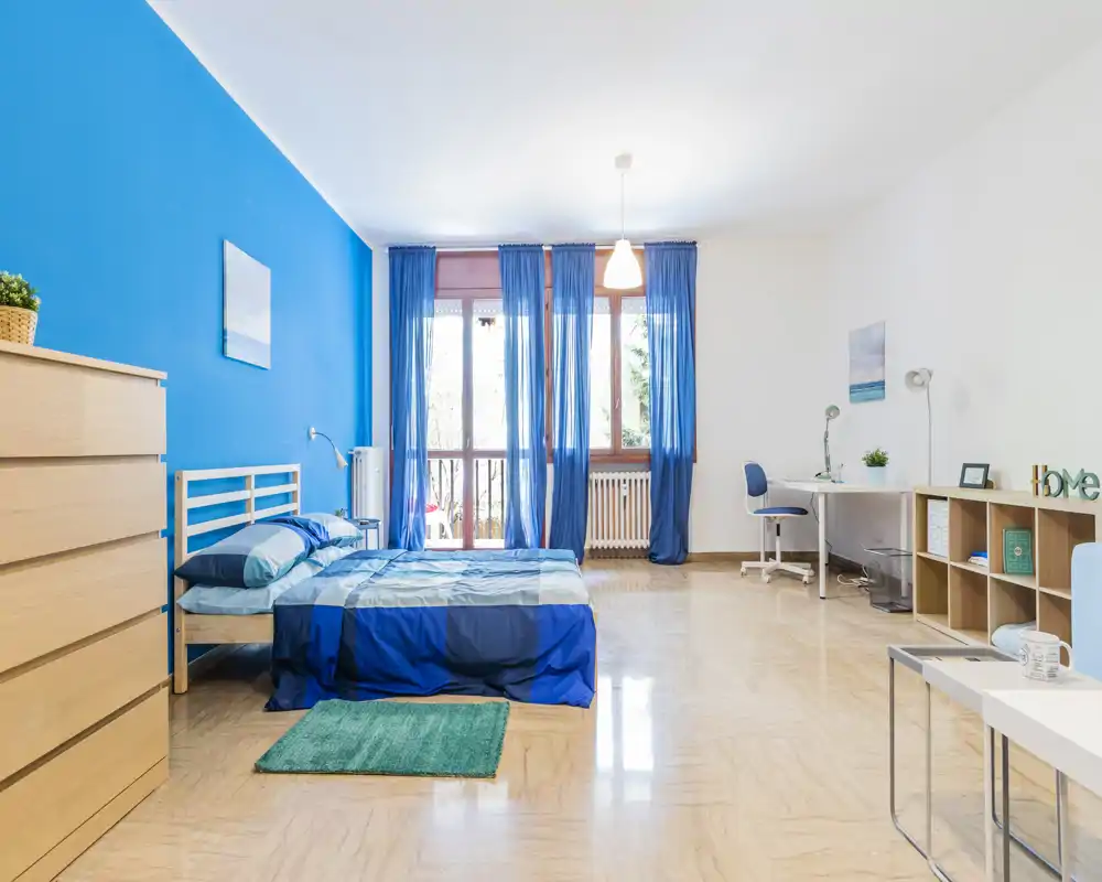Bright, spacious bedroom with a double bed dressed in blue linens, large window with sheer blue curtains and a small workspace — ideal for a comfortable stay.