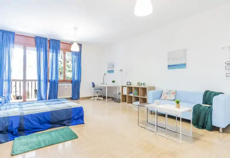 Bright, airy bedroom-studio with a blue-themed single bed by large windows, a compact desk area and a light sofa — ideal for a single tenant or student looking for a fresh, modern space.