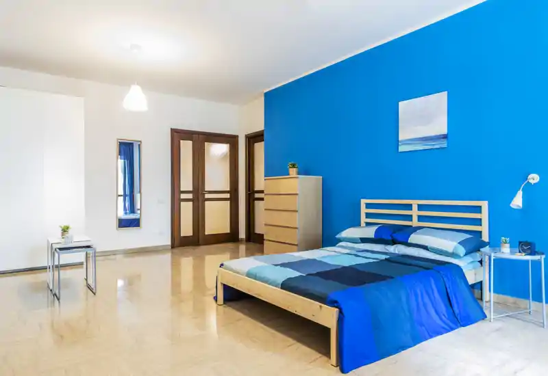 Bright, modern bedroom with a wooden double bed against a vivid blue accent wall, matching bedding and minimal, functional furniture that creates a fresh and welcoming space.