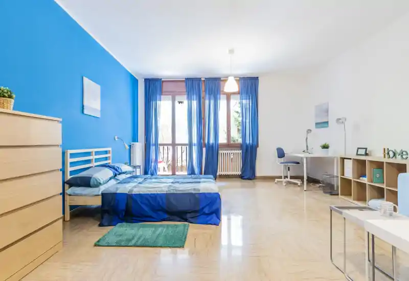 Bright, spacious bedroom with a double bed dressed in blue linens, large window with sheer blue curtains and a small workspace — ideal for a comfortable stay.