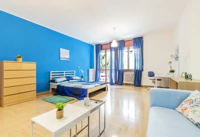 Spacious, bright bedroom with a double bed, wooden flooring and large windows letting in abundant natural light. The room is styled in blue tones with a comfortable seating area, desk and storage chest—ideal for a cozy, modern rental.