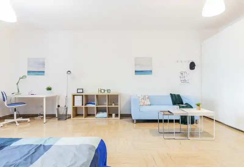 Bright, modern studio bedroom with a visible bed in the foreground, a light-blue sofa and compact desk—ideal for a single occupant or couple seeking a clean, airy space.