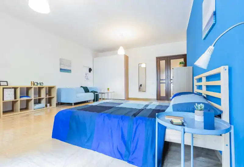 Bright, modern bedroom with blue-accented bedding and a wooden bed frame; a small round bedside table and a sofa add comfort and practical seating.