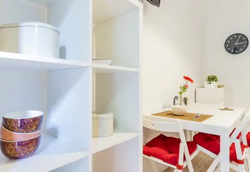 Bright, compact dining area with a white table, folding chairs topped with red cushions, a decorative placemat and a small vase with flowers — ideal for cozy meals.