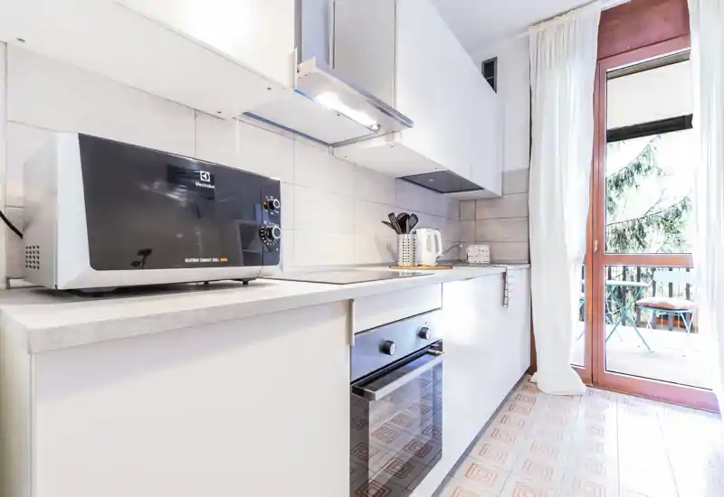 Bright modern kitchenette with white cabinets, countertop appliances (microwave, kettle) and an oven. A glass door opens to a small balcony, bringing natural light and a welcoming atmosphere.