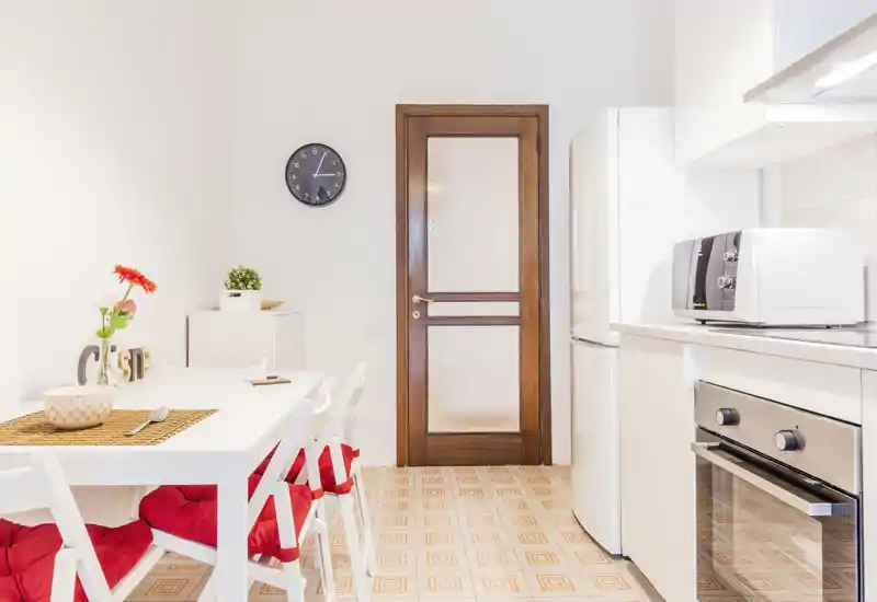 Bright, compact kitchen with integrated dining table, white cabinetry, oven and fridge — cozy red seat cushions and a floral centerpiece add warmth.