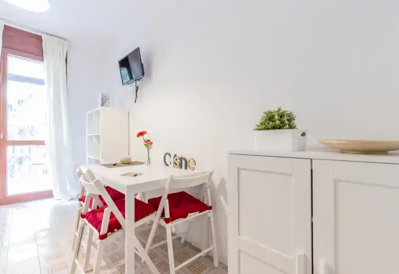 Small bright dining area with a white table and four folding chairs topped with red cushions, decorative flowers and shelving beside a glass door letting in natural light.