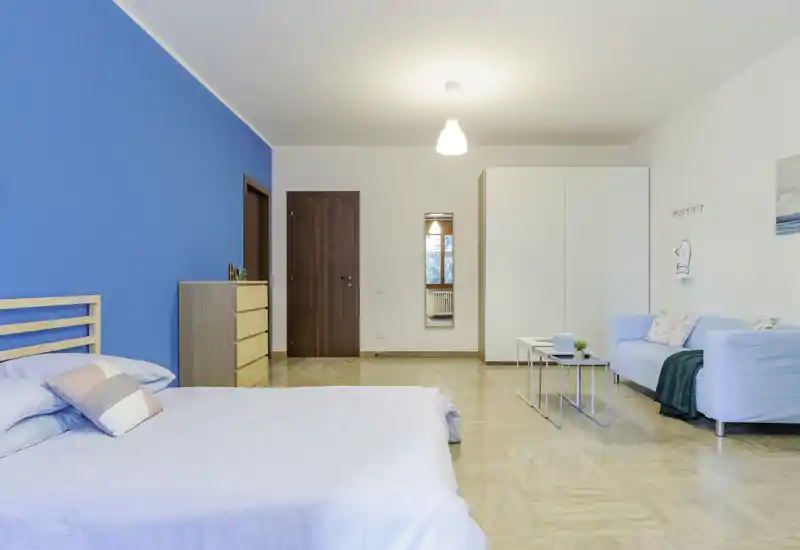 Spacious, bright bedroom with a light wooden bed and pastel bedding. The room features a bold blue accent wall, a dresser, wardrobe and a small seating area, offering a calm, modern stay.