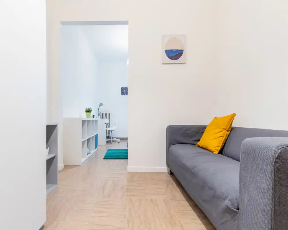 Bright, minimal living area with a grey sofa and a yellow cushion; view into an adjacent room with shelving and a small desk, perfect for showcasing a compact apartment living space.