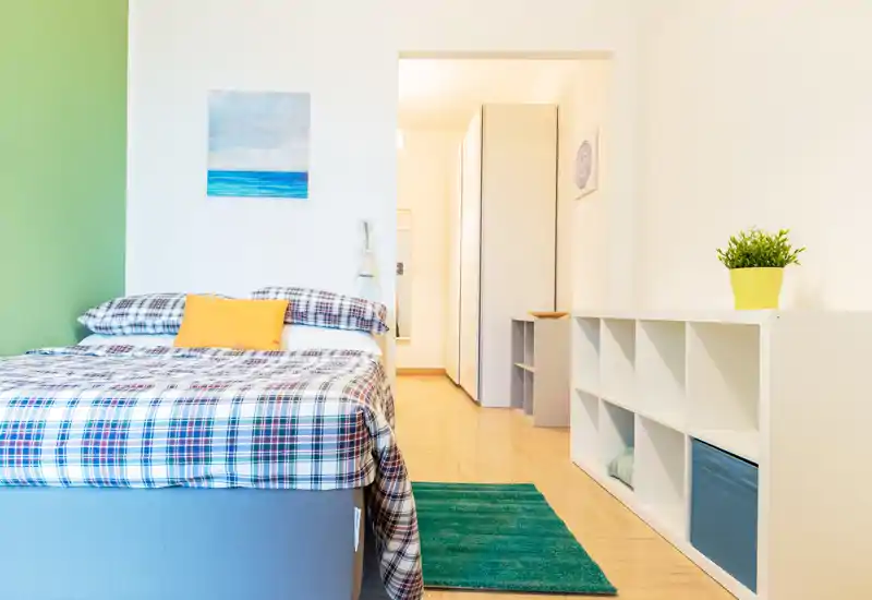 Bright, modern bedroom with a neatly made double bed in checked linens, a pop of yellow on the pillow, a green accent wall and organized shelving—ideal for rental photos highlighting comfort and clean styling.