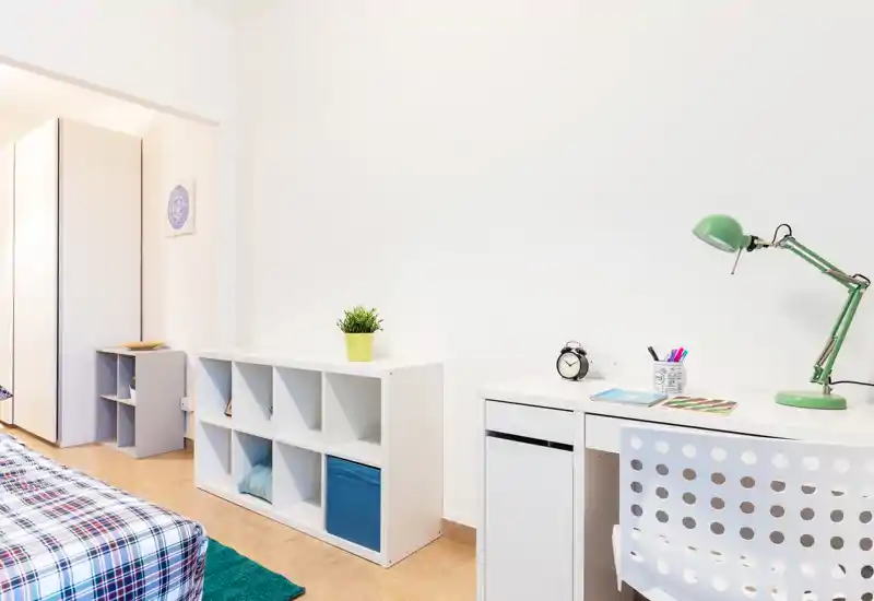 Bright, modern bedroom corner with a bed in checked bedding, organized storage cubes and a white desk with a green task lamp — minimal and functional decor ideal for a rental listing.