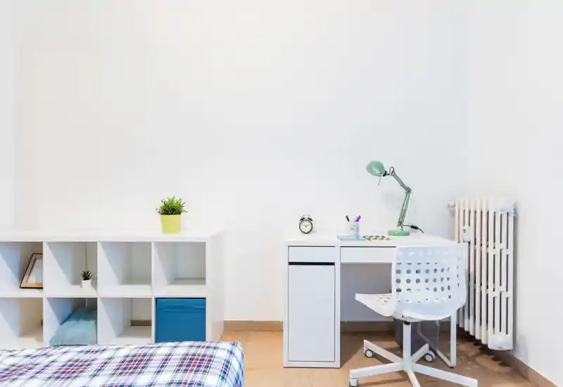 Bright, minimalist bedroom corner with a single bed partially visible, a compact white desk with a green task lamp and a perforated chair — ideal for a student or home office setup.