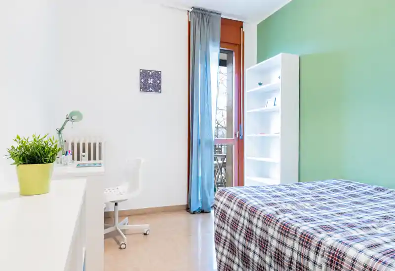 Bright, neatly styled bedroom with a patterned bedspread, a work desk and shelving by a balcony door; fresh green accent wall and natural light create a welcoming atmosphere.