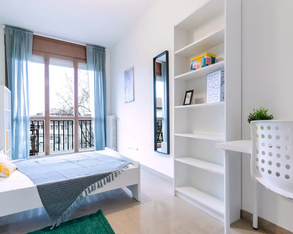 Bright, modern bedroom with a white bed, soft blue textiles, a shelving unit and large window leading to a balcony that floods the room with natural light.