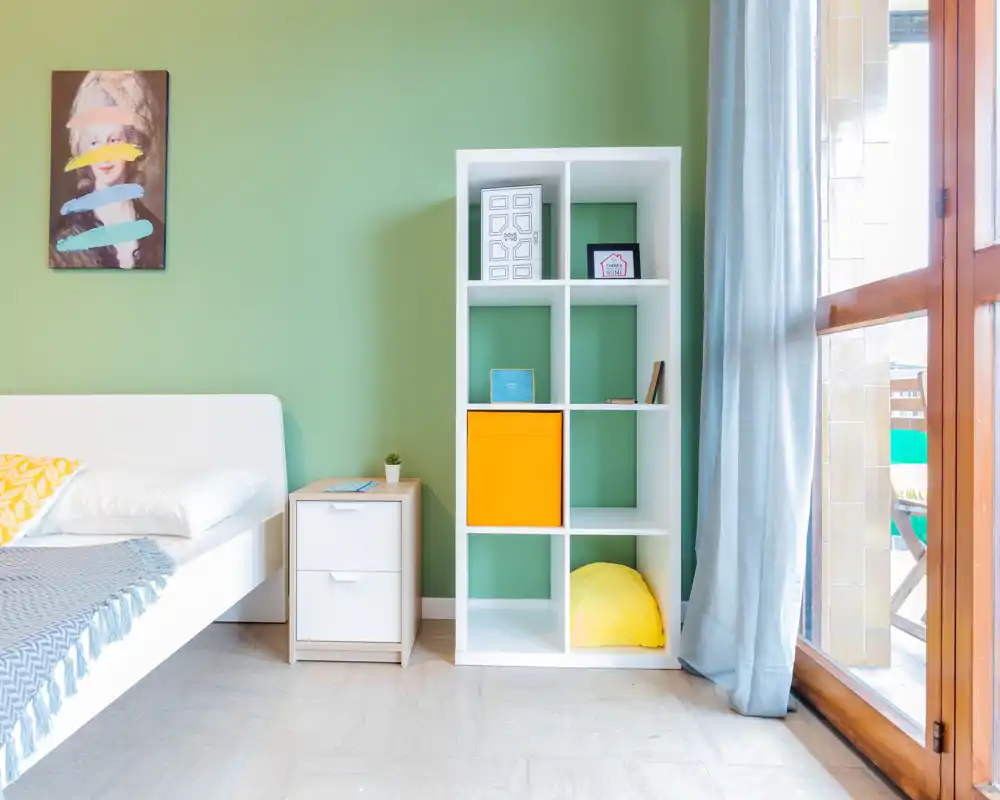 Bright, modern bedroom with a white bed, bedside drawer and a cubed shelving unit against a soft green wall; large window/door brings abundant natural light and a calm, airy feel.
