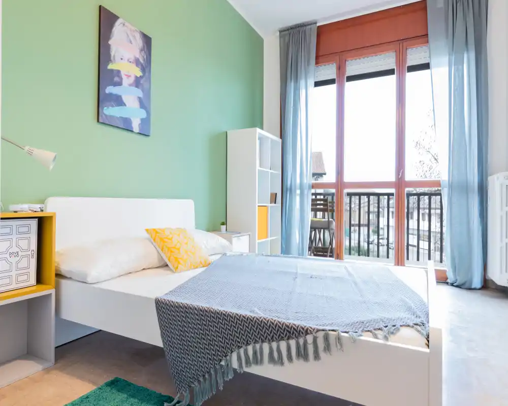 Bright, modern bedroom with a white platform bed, colorful cushions and large floor-to-ceiling window letting in natural light; tasteful green accent wall and shelving create a fresh, inviting feel.