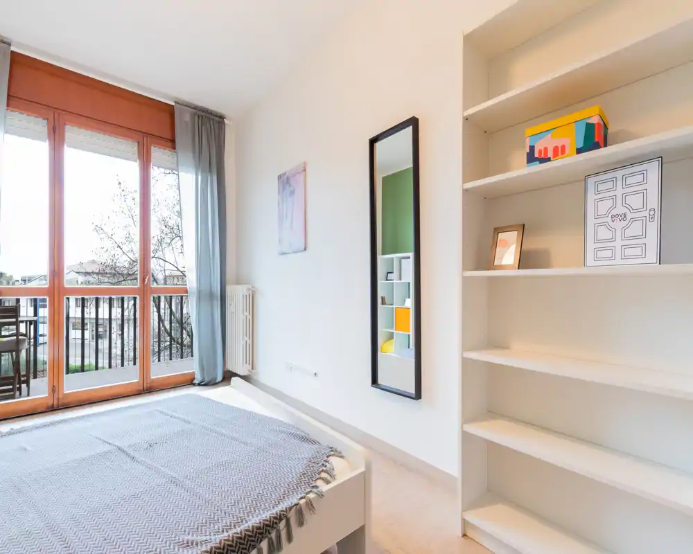 Bright, minimalist bedroom with a large glass door to the balcony, simple bed frame and open shelving—ideal for a rental listing showing natural light and neat staging.
